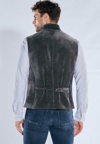 Graue Cordweste mit hohem Kragen, rückseitigem Gürtel-Detail und glatter Textur, kombiniert mit einem gestreiften Langarmshirt.