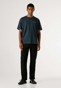 Tamnoplava kratka majica s okruglim izrezom, u kombinaciji s crnim hlačama i crnim cipelama. Tkanina izgleda mekano i blago teksturirano.