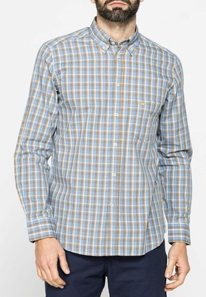 Uomo che indossa una camicia a quadri blu, bianca e beige con colletto abbottonato e pantaloni blu navy, in piedi su sfondo bianco.