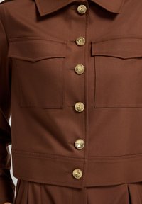 Brun jacka med strukturerade lockfickor på bröstet och gyllene metallknappar framtill, i kombination med matchande plisserade byxor.