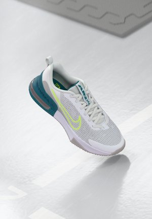 Hvid og teal atletiksko med neon-gult swoosh-logo og mesh-overdel, vist flydende over lys grå tekstureret gulv.