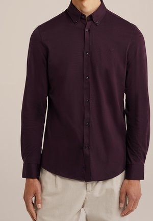 Man draagt een nauwsluitend, donkerpaars overhemd met lange mouwen en beige broek tegen een neutrale achtergrond.