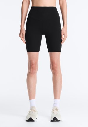 Osoba ubrana w czarne dopasowane szorty z wysokim stanem, białe skarpety za kostkę i jasne buty do biegania, stojąca na jednolitym białym tle.