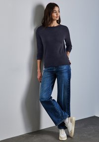 Mörkblå långärmad topp med rund halsringning, matchad med vida blå jeans. Vita sneakers och en slät texturerad vägg i bakgrunden.