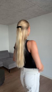 Lunghi capelli biondi lisci raccolti con una clip nera. La persona indossa una canotta nera e pantaloni bianchi a vita alta, con una stanza minimalista sullo sfondo.