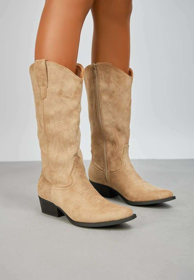 Bottines de cowboy en suède beige avec des orteils pointus, des coutures décoratives et un petit talon carré. Texture lisse avec une finition polie.