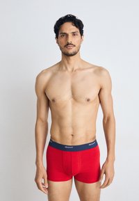 Rote Baumwoll-Boxershorts mit einem marineblauen Bund, der das "GANT"-Logo zeigt, enganliegendes Design und glatte Textur. Das Modell steht vor einem schlichten Hintergrund.