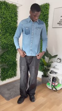 Uomo che regola i pantaloni cargo neri, indossando una camicia di jeans blu e scarpe nere, in piedi all'interno vicino a una pianta verde e a un tavolino rotondo con una macchina fotografica.