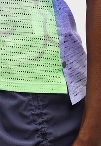 Κοντινό πλάνο αθλητικού ρουχισμού με πράσινο και μωβ αναπνεύσιμο τοπ από δίχτυ και σκούρο μπλε σορτς στο πλευρό και το χέρι ενός ατόμου.