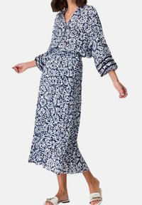 Femme portant une robe midi à motifs floraux blancs et bleu marine avec des manches 3/4 et des sandales blanches à enfiler avec des accents de chaîne dorée.
