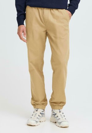 Beige jogger pants with elastic waistband and cuffs, featuring a smooth texture and side pockets, paired with white and gray athletic shoes.