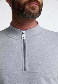 Grauer Pullover mit hohem Kragen, ausgestattet mit einem Reißverschluss vorne und strukturiertem Stoff. Auffälliges Detail: Metall-Reißverschlussbeschläge.