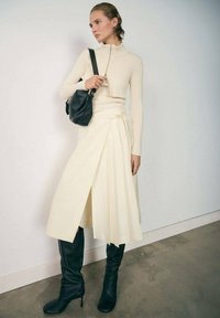Knit cream top with a zipped high collar and long sleeves. Flowing white wrap skirt, paired with fitted black knee-high boots and a black bag.