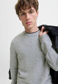 Un pull en tricot gris avec un col rond et des poignets côtelés, porté par un mannequin tenant une veste en cuir noire sur une épaule.
