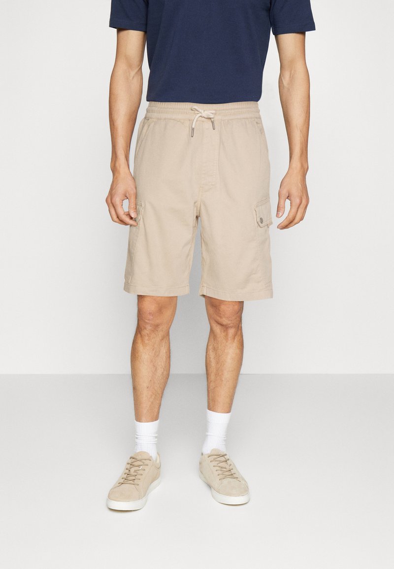 Shorts cargo beiges avec une taille élastique, des poches latérales, et une coupe décontractée, associés à un t-shirt bleu marine et des baskets beige clair.
