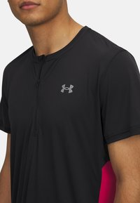 Musta urheilupaita, jossa on osittainen vetskari, lyhyet hihat ja vaaleanpunaiset sivuakselit. Kankaan pinta on teksturoitu, ja rinnassa on Under Armour -logo.