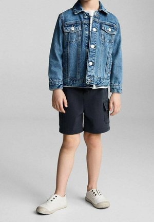 Bambino con giacca di jeans blu, pantaloni cargo neri e sneakers bianche senza lacci, in piedi su uno sfondo grigio semplice.