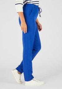 Blauwe, taps toelopende broek met zijzakken, een trekkoord in de taille en een gladde textuur. Draag met witte sneakers met een gestructureerd ontwerp.