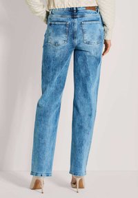 Persoon in lichtblauwe rechte jeans en beige hoge hakken, gezien van achter tegen een egale lichte achtergrond.