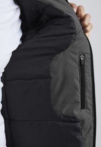 Intérieur d'une veste en duvet grise, avec doublure noire, texture matelassée et une poche zippée sur le côté droit. Fini lisse et design moderne.