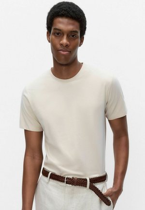 Jeune homme aux cheveux courts et bouclés portant un t-shirt beige, un pantalon clair et une ceinture tressée marron, une main dans la poche.