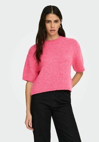 Femme posant dans un pull en maille à manches courtes rose vif et un pantalon noir, sur un fond clair uni.