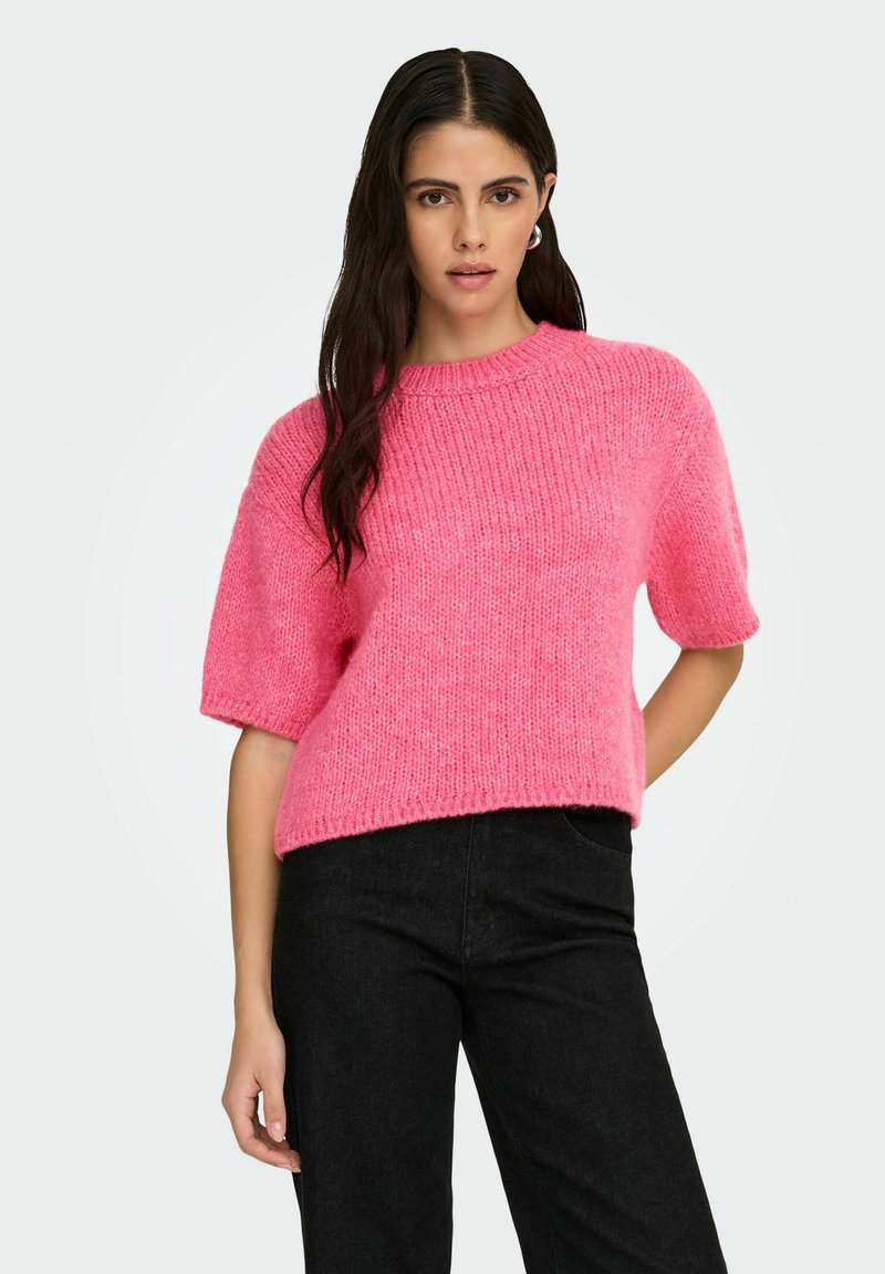 Femme posant dans un pull en maille à manches courtes rose vif et un pantalon noir, sur un fond clair uni.