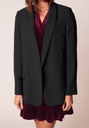 Kvinde iført en sort blazer over en burgunderfarvet velour slå-om-kjole, stående foran en lys beige baggrund.