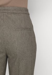 Gros plan sur une poche de pantalon texturée marron et beige avec une ceinture ajustée, portée avec un haut côtelé marron.