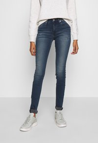 Persona que lleva jeans skinny azul oscuro con los bajos doblados, zapatillas blancas y una camiseta de manga larga de color claro con motas, de pie contra un fondo liso.