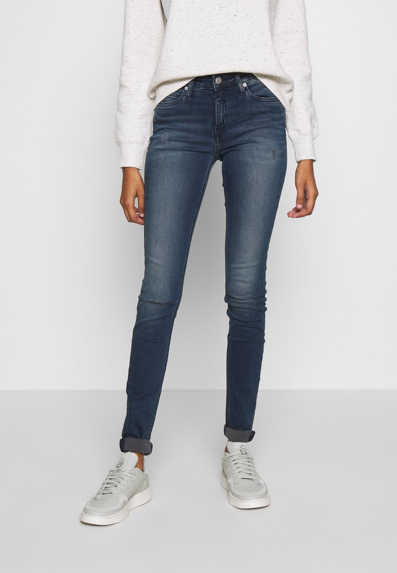 Persona que lleva jeans skinny azul oscuro con los bajos doblados, zapatillas blancas y una camiseta de manga larga de color claro con motas, de pie contra un fondo liso.