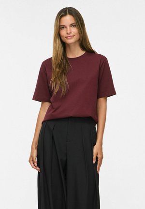 Giovane donna con lunghi capelli castani che indossa una maglietta bordeaux a maniche corte e pantaloni neri a vita alta plissettati, in piedi su uno sfondo bianco.