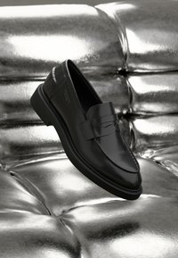 Mocassins en cuir noir au design épuré, avec des accents cousus et un bout rond, reposant sur une surface argentée rembourrée.
