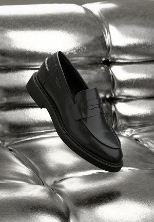 Mustad mustad nahkadega penny loafers, elegantse disainiga, õmmeldud aktsentidega ja ümarate varvastega, puhkamas hõbedasel pehmendatud pinnal.