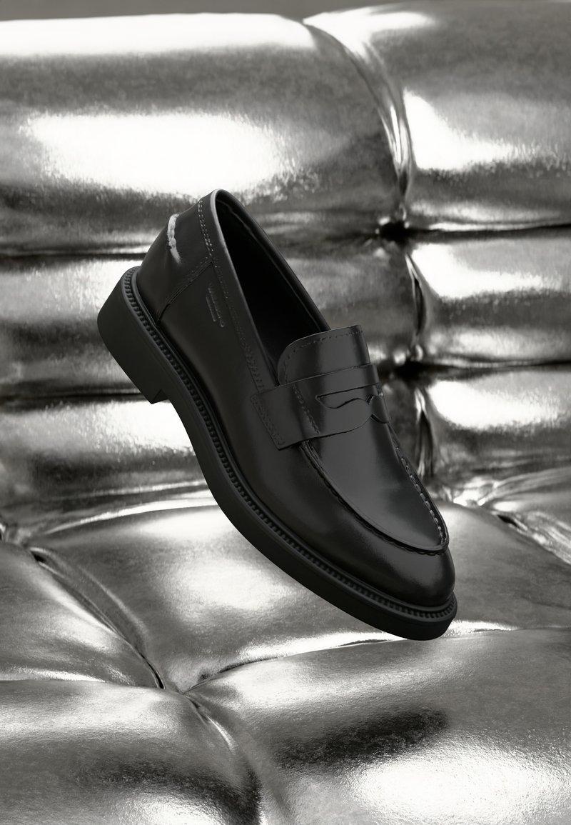 Sorte skinn penny loafers med et slankt design, sydde detaljer og rund tå, som hviler over en sølvfarget polstret overflate.