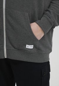 Sudadera con cremallera gris, con puños y dobladillo de canalé. Presenta dos bolsillos frontales y una pequeña etiqueta de marca cerca del dobladillo. Textura de tela suave.