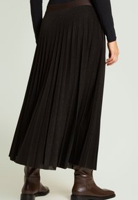 Falda maxi negra plisada con acabado texturizado, con una cinturilla ancha. Combinada con botas marrones hasta el tobillo para un look en capas.