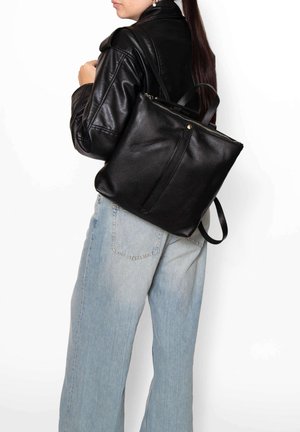 Femme en veste en cuir noire et jean bleu clair portant un sac à dos en cuir noir sur une épaule, vue de profil.