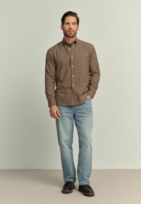 Camisa de botones a cuadros marrones con mangas largas, combinada con jeans azul claro y zapatos oscuros, con un fondo neutro.