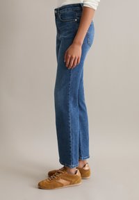 Person wearing blue straight-leg jeans and brown suede sneakers standing against a neutral background.