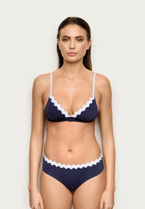 Donna che indossa un bikini blu navy con bordi bianchi smerlati, in piedi davanti a uno sfondo chiaro e uniforme, rivolta in avanti con espressione neutra.