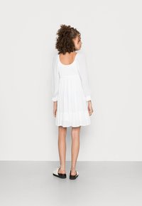 Vestido branco até ao joelho com corpete ajustado, mangas bufantes e saia em camadas e rodada; combinado com sapatos de slip-on em tons claros.