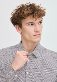 Camisa de botones con patrón en marrón y blanco con pequeños diseños florales, con cuello extendido y mangas largas. Textura de tela suave.