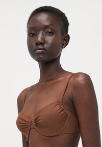 Brun moldet bikini-overdel med justerbare stropper og samlet detalj i midten. Materialet ser glatt og elastisk ut.