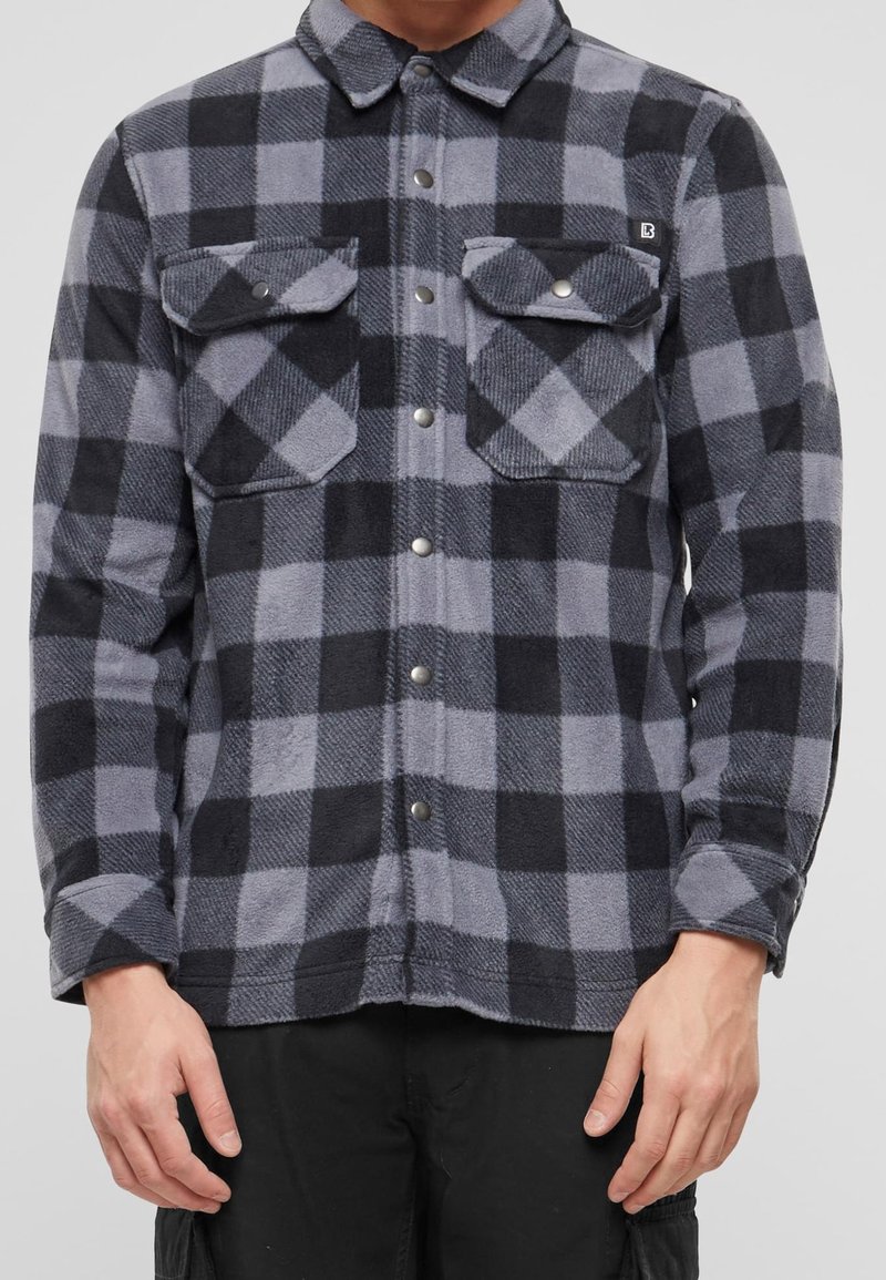 Homme portant une chemise à carreaux grise et noire à boutons, avec deux poches poitrine et manches longues, associée à un pantalon noir.
