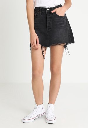 Personne portant une mini-jupe en jean noir effilochée, un T-shirt blanc et des baskets montantes blanches sur un fond blanc uni.