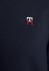 Donkerblauwe stof met geborduurde witte en rode verweven letters "T" en "H" in het midden van het borstgebied.