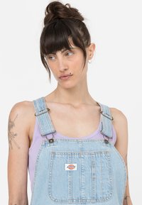 Pettorina in denim di colore azzurro chiaro con spalline regolabili, tasca frontale e patch con logo. Sotto si indossa una canotta lavanda.