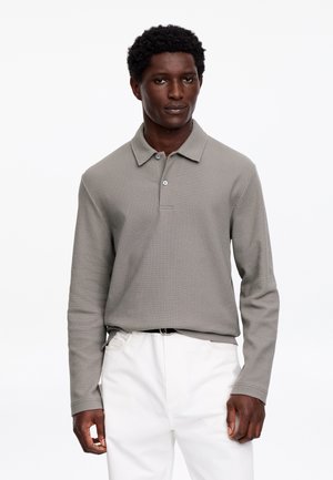 Uomo che indossa una polo a maniche lunghe grigia infilata in pantaloni bianchi, in piedi contro uno sfondo bianco uniforme.