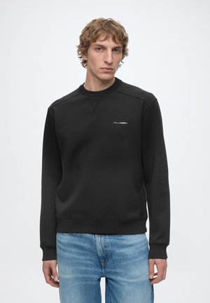 Man wearing a plain black sweatshirt with small white logo on chest and blue jeans, standing against a plain light background.